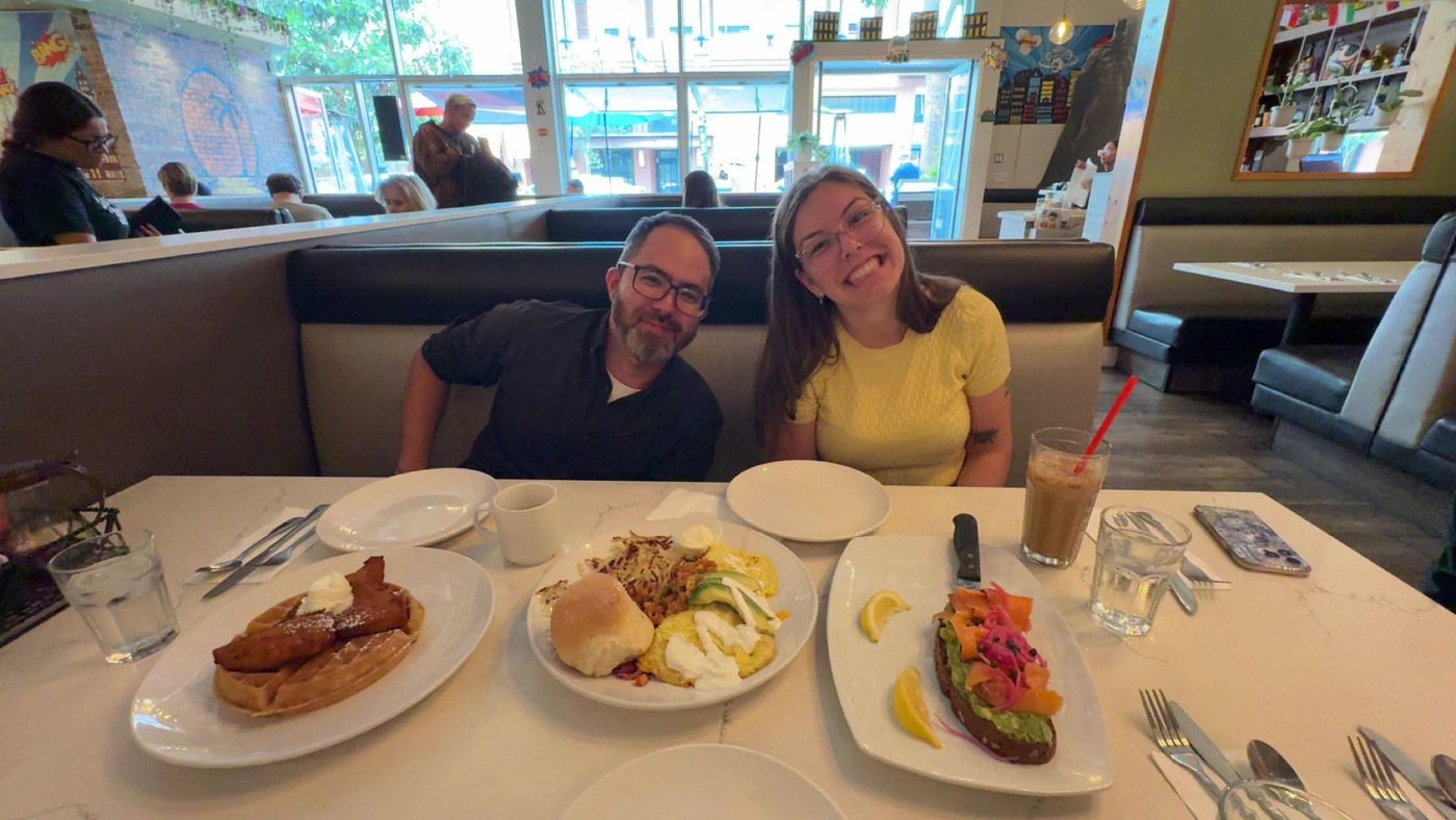 Anna in a yellow top and her friend Marc at a brunch table laid with avocado toast, eggs, and coffee.