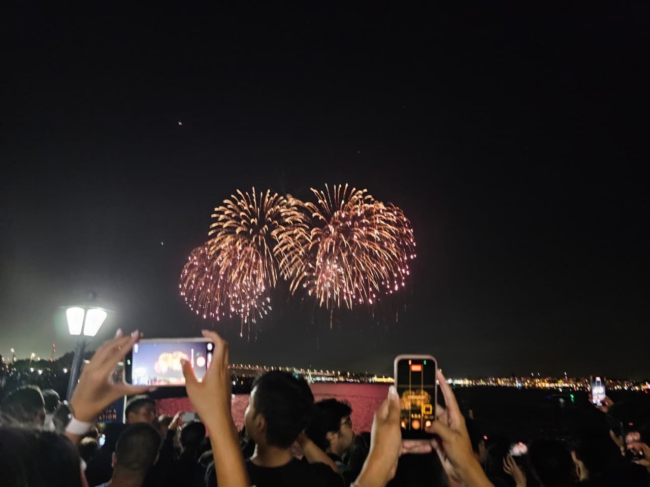 Fireworks bursting over San Diego Bay as someone films on their phone.