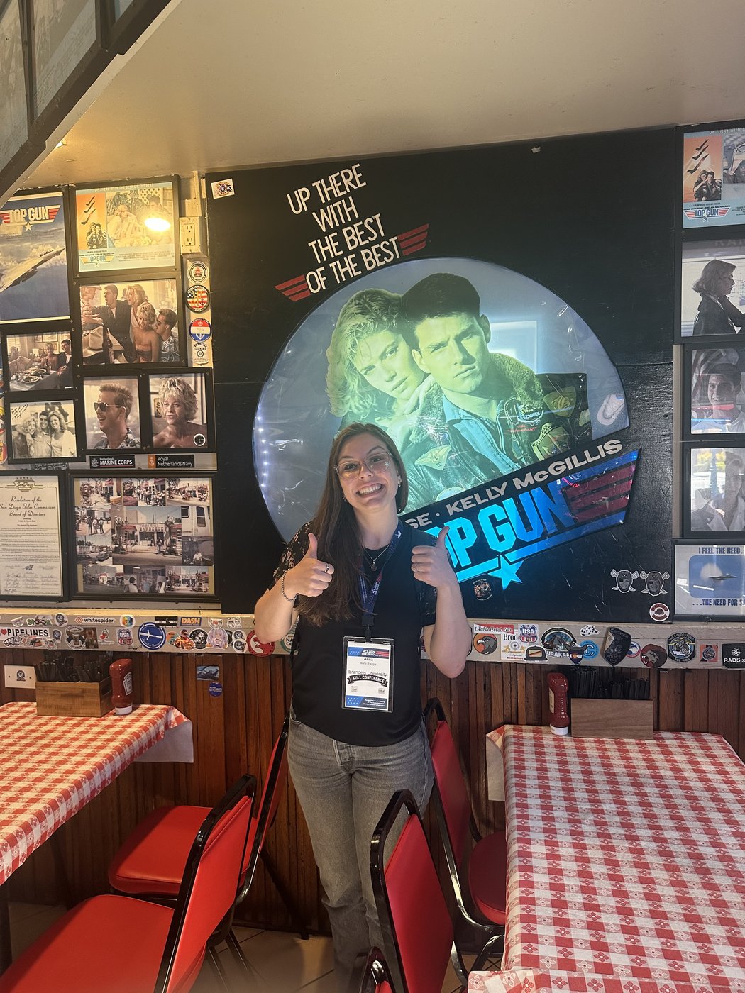 Anna at a red-checkered table with Top Gun projected on the wall beneath a sign reading 'up here we're the best of the best.'