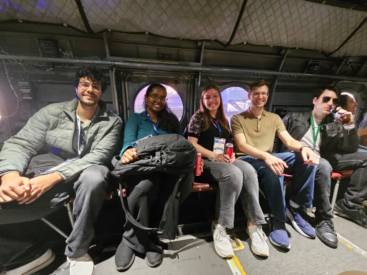 Five friends squeezed onto the bench seats inside the cabin of a tactical helicopter aboard the USS Midway, round porthole windows behind them.