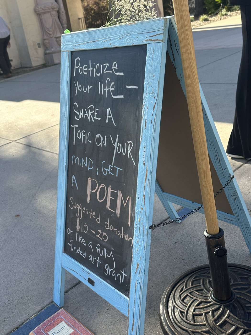 A pale-blue A-frame chalkboard sign reading 'Poeticize your life — share a topic on your mind, get a poem. Suggested donation $10 to 20, or like a fully funded art grant.'