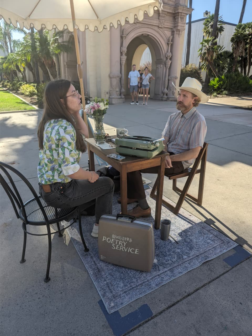 A street poet at a small table under a stone archway in Balboa Park, composing a poem on a typewriter.