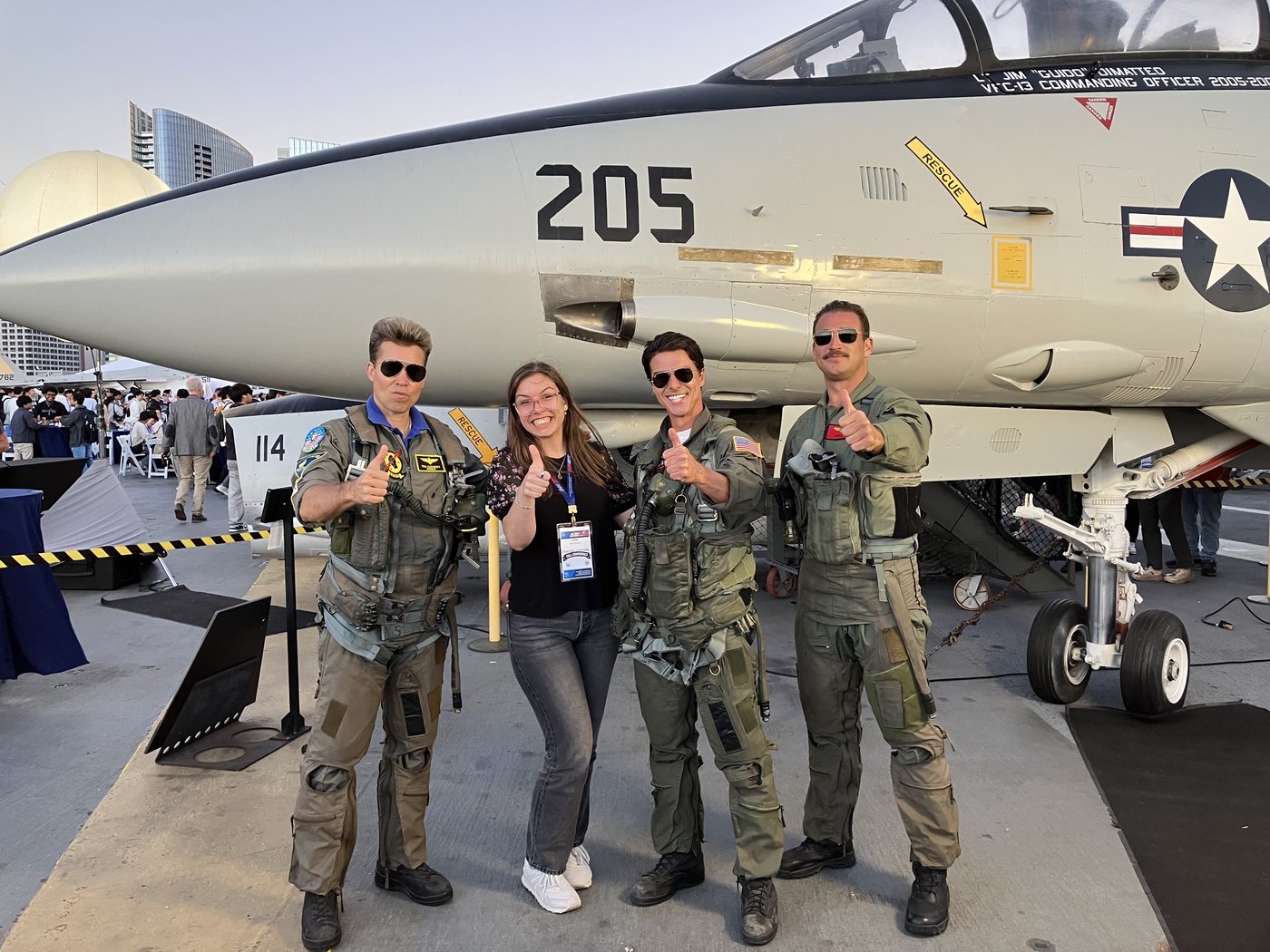Four friends in green flight suits striking the Top Gun finger-gun pose in front of a fighter jet numbered 205.