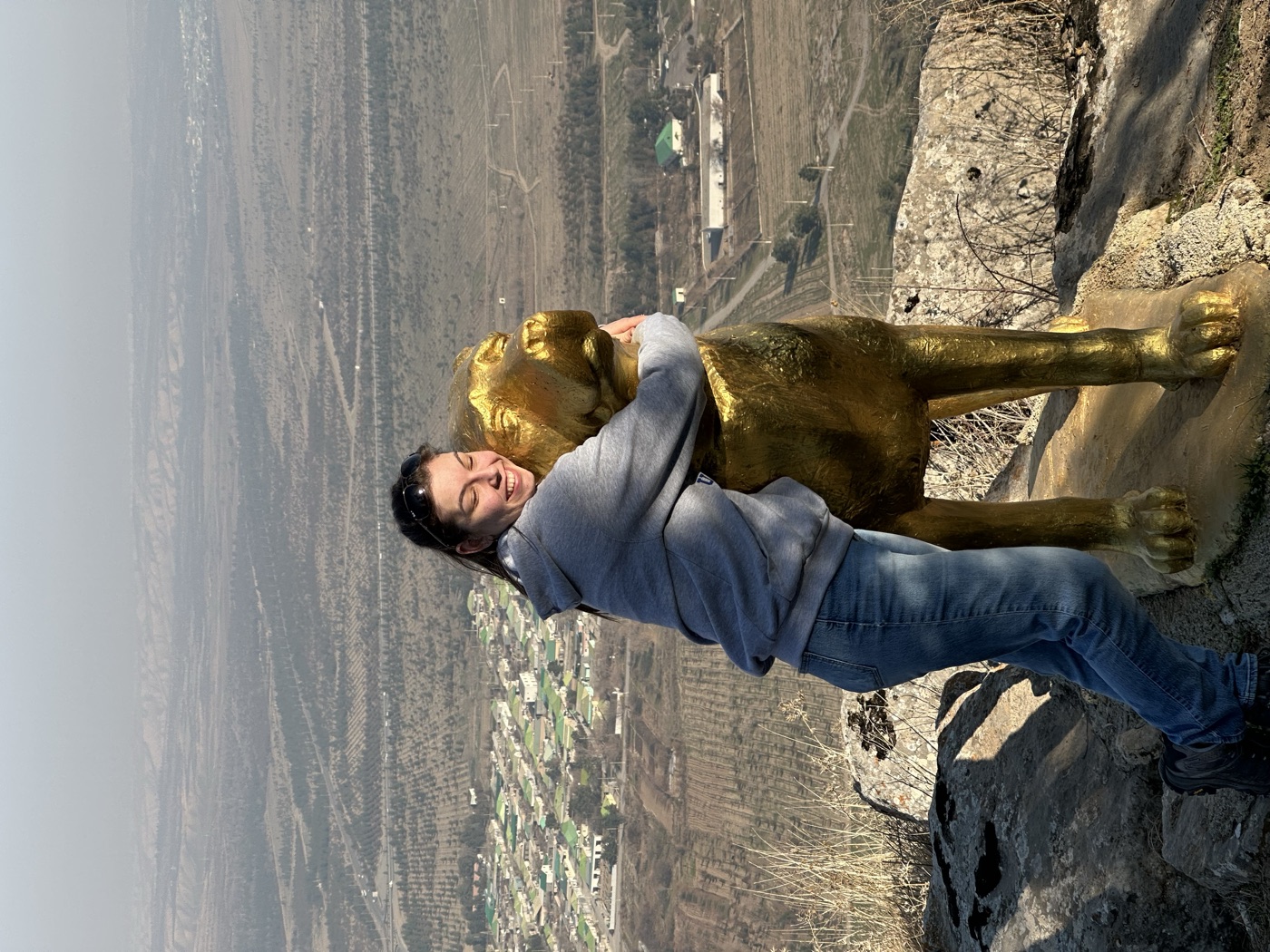 Anna hugging a golden statue of an Alabai dog on a hilltop overlooking a valley.