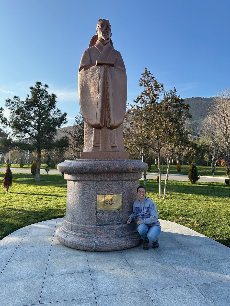 The author crouching and smiling beside a tall bronze-colored statue of Du Fu in Ashgabat, Turkmenistan, on a granite pedestal; the plaque reads Du Fu 712–770.