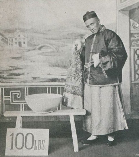 Ching Ling Foo in a long robe and cap on stage beside a large bowl and a sign reading 100 LBS. Public domain, via Wikimedia Commons.