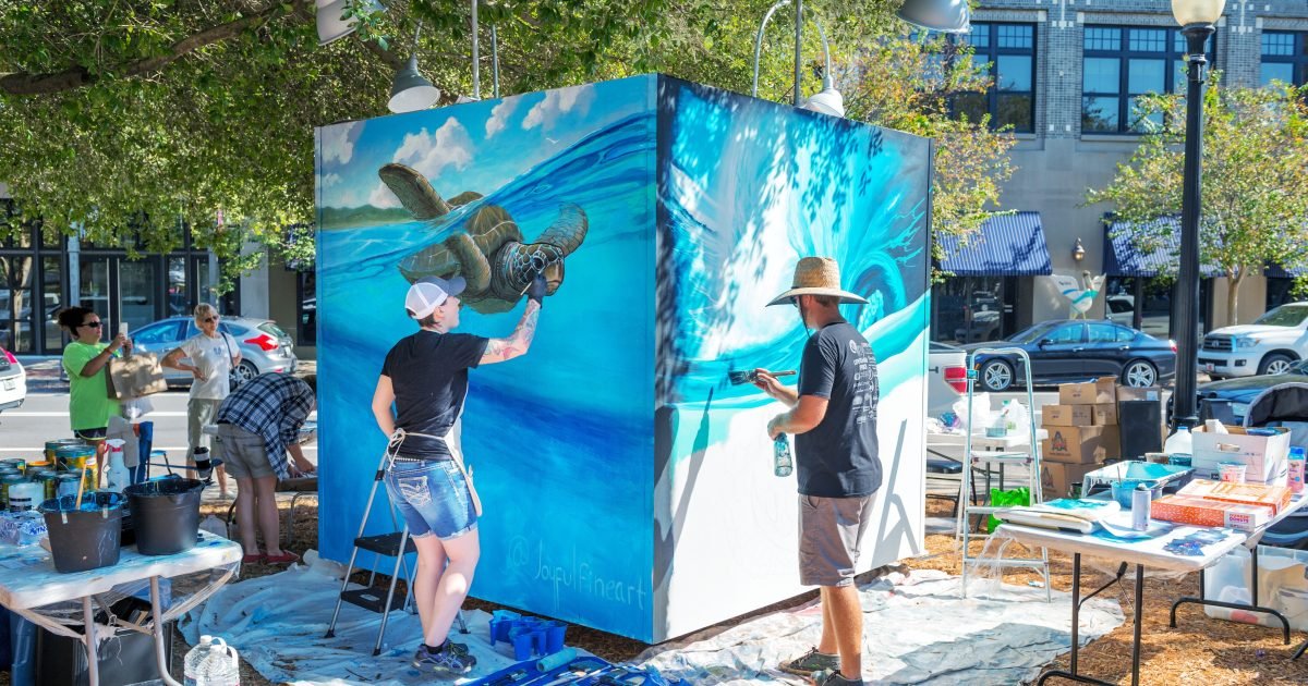 Two artists painting a sea turtle and a curling blue wave on a large outdoor art cube in downtown Pensacola during Foo Foo Festival.