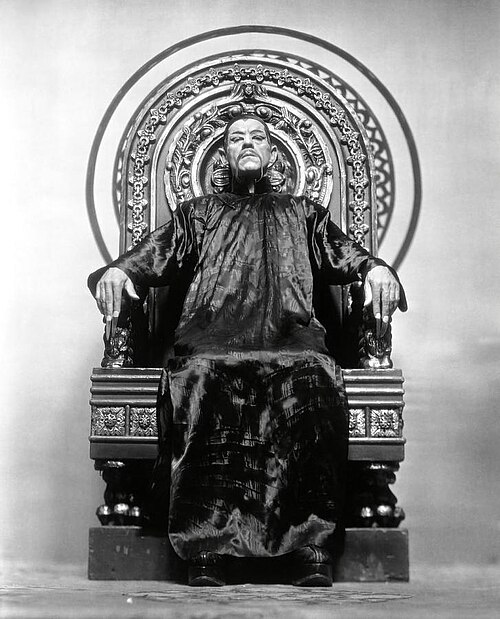 Boris Karloff as Fu Manchu, seated on an ornate throne in a dark silk robe, in The Mask of Fu Manchu (1932). Via Wikimedia Commons.