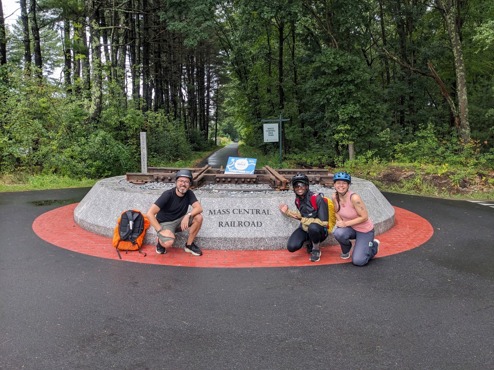 Marc, Anna, and Sharde crouch smiling in front of a Mass Central Railroad monument made of rail track, bikes and helmets on, pine woods behind.