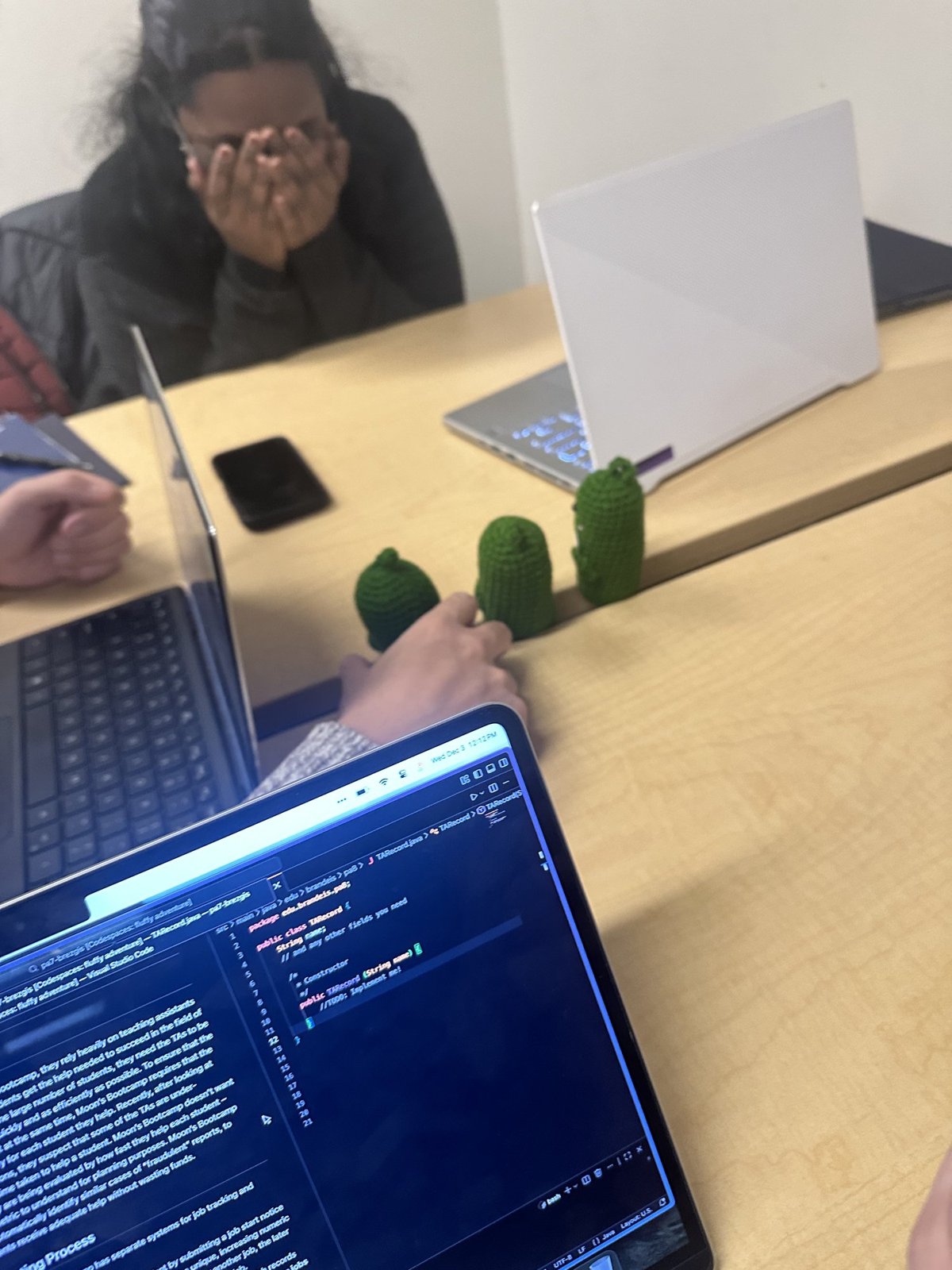 A friend at a library table hiding her face behind her hands, laughing, with two small crocheted pickles standing on the table beside a laptop.