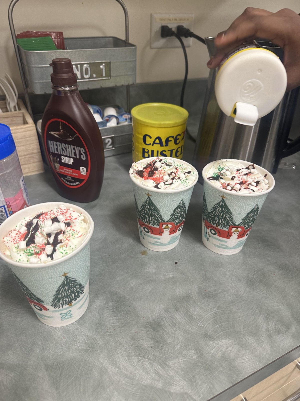 Three paper cups of coffee topped with whipped cream and chocolate drizzle on a counter, a bottle of Hershey's syrup beside them, a hand pouring milk from a thermos.