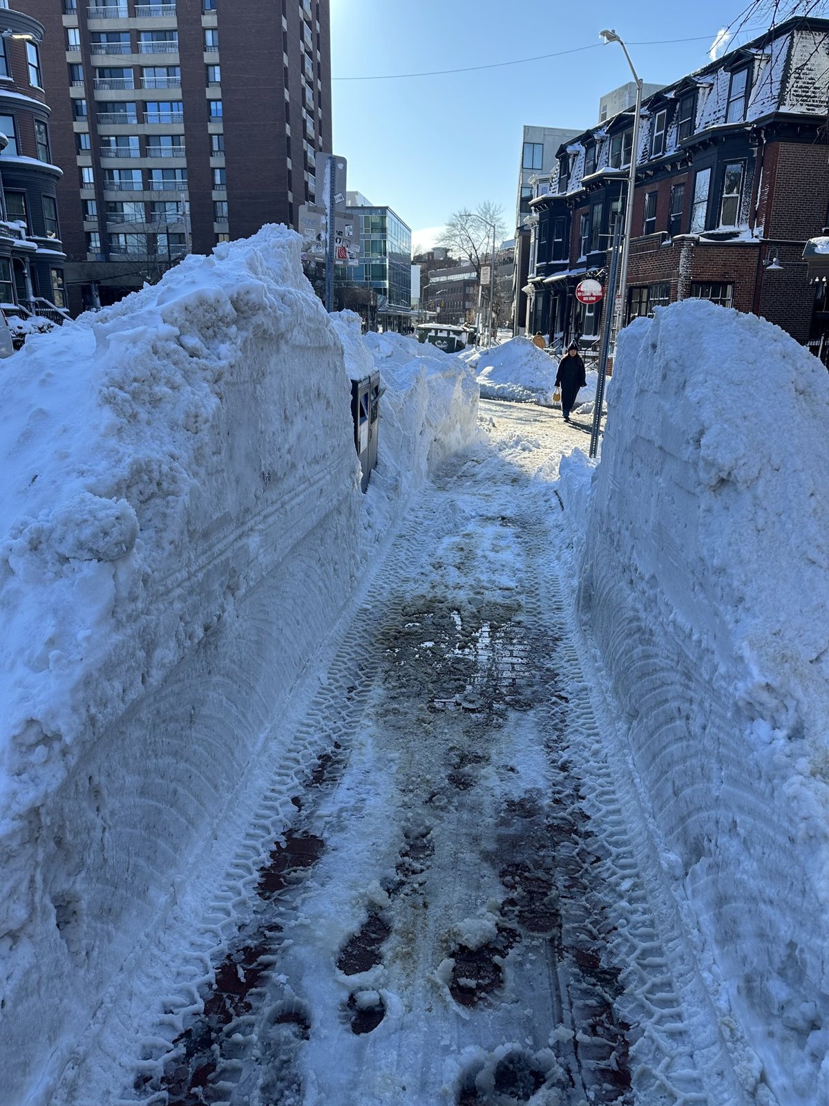 A narrow sidewalk path cut between two waist-high walls of plowed snow on a Cambridge street, apartment buildings ahead.