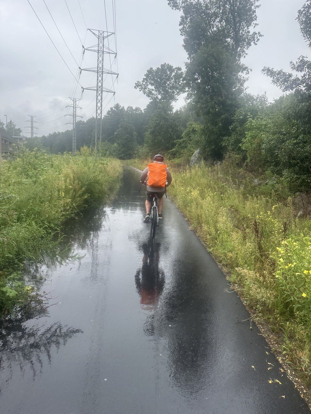 A cyclist in an orange rain cover rides away down a flooded rail trail under grey skies, transmission towers ahead.