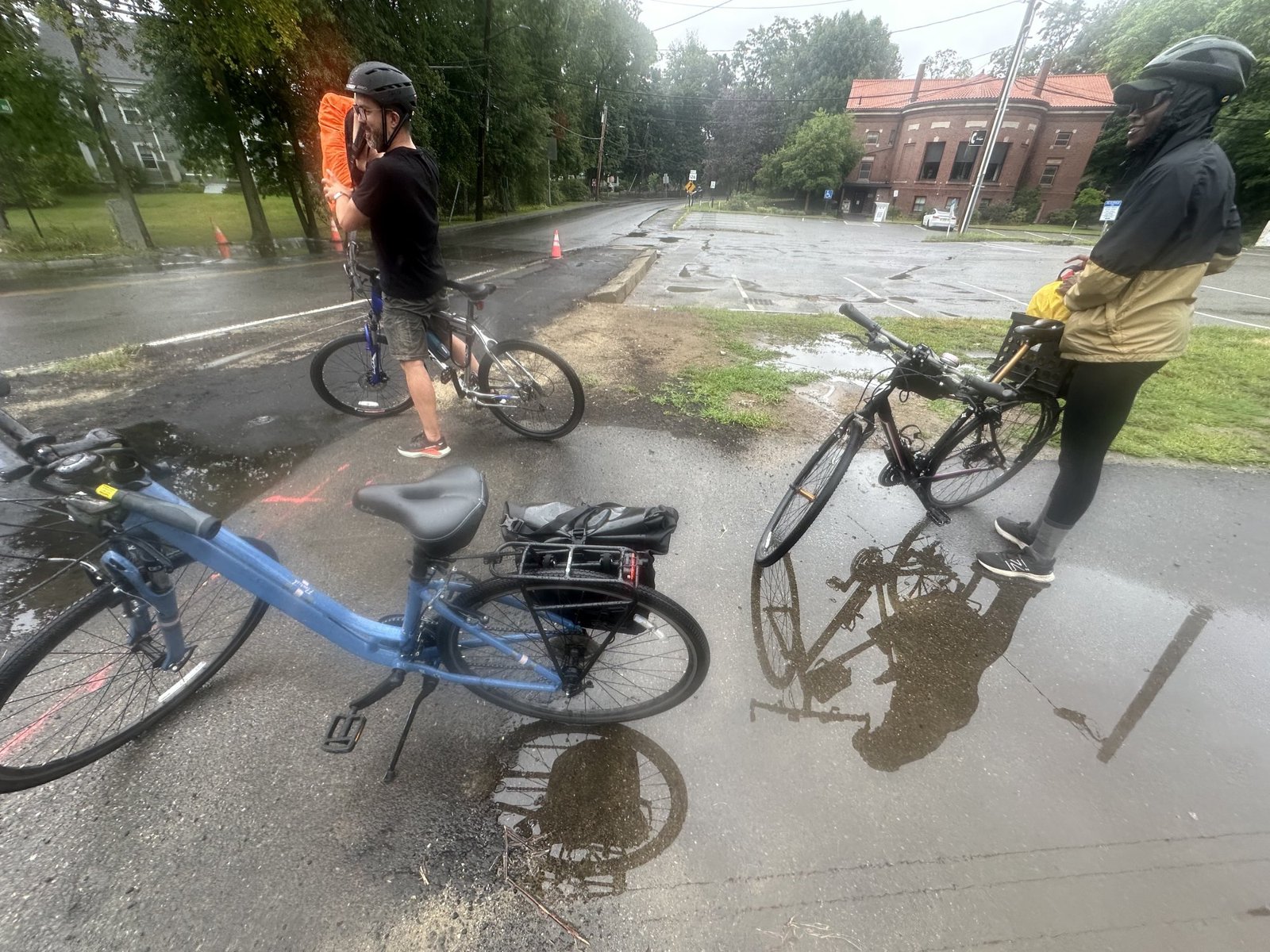 Marc and Sharde standing with their bikes at a wet intersection, a blue bike in the foreground, a brick former train station across the street.