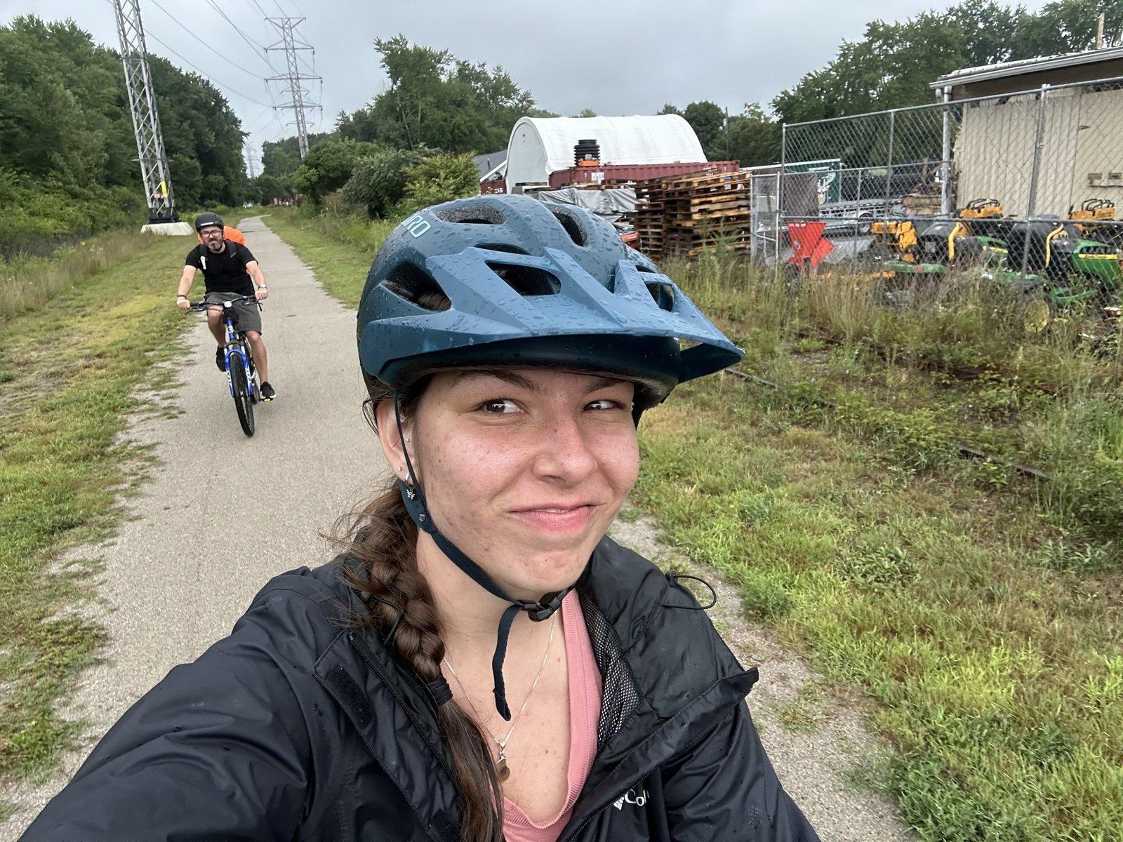 Anna in a bike helmet takes a selfie on the trail, a braid over her shoulder, with two friends biking toward her in the background.