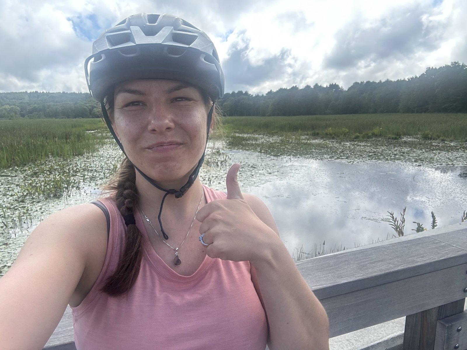 Anna in a helmet gives a thumbs up on a boardwalk over a wide marsh under cloudy skies.