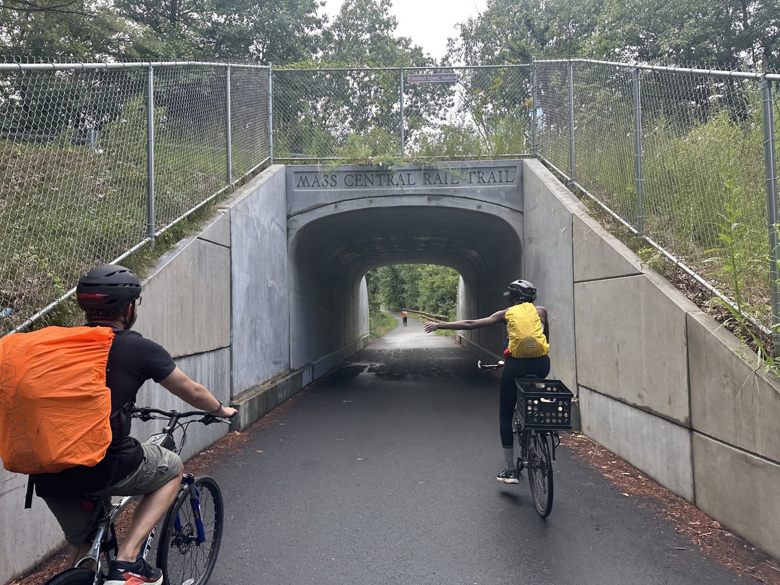 Two cyclists ride into a concrete underpass labeled Mass Central Rail Trail, one with an orange pack, one with a yellow one.
